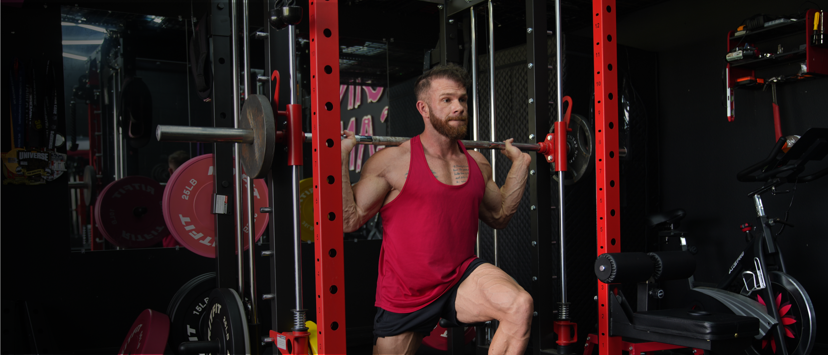 A man in a red tank top and black shorts performing a squat inside a red power rack in a gym setting.