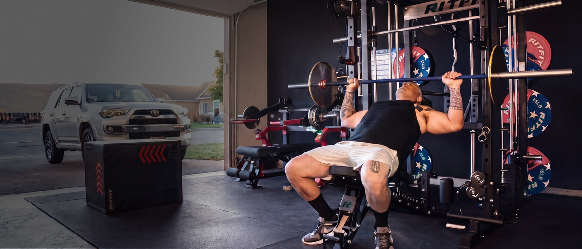Man performing bench press on RitFit power rack in home garage gym with weight plates