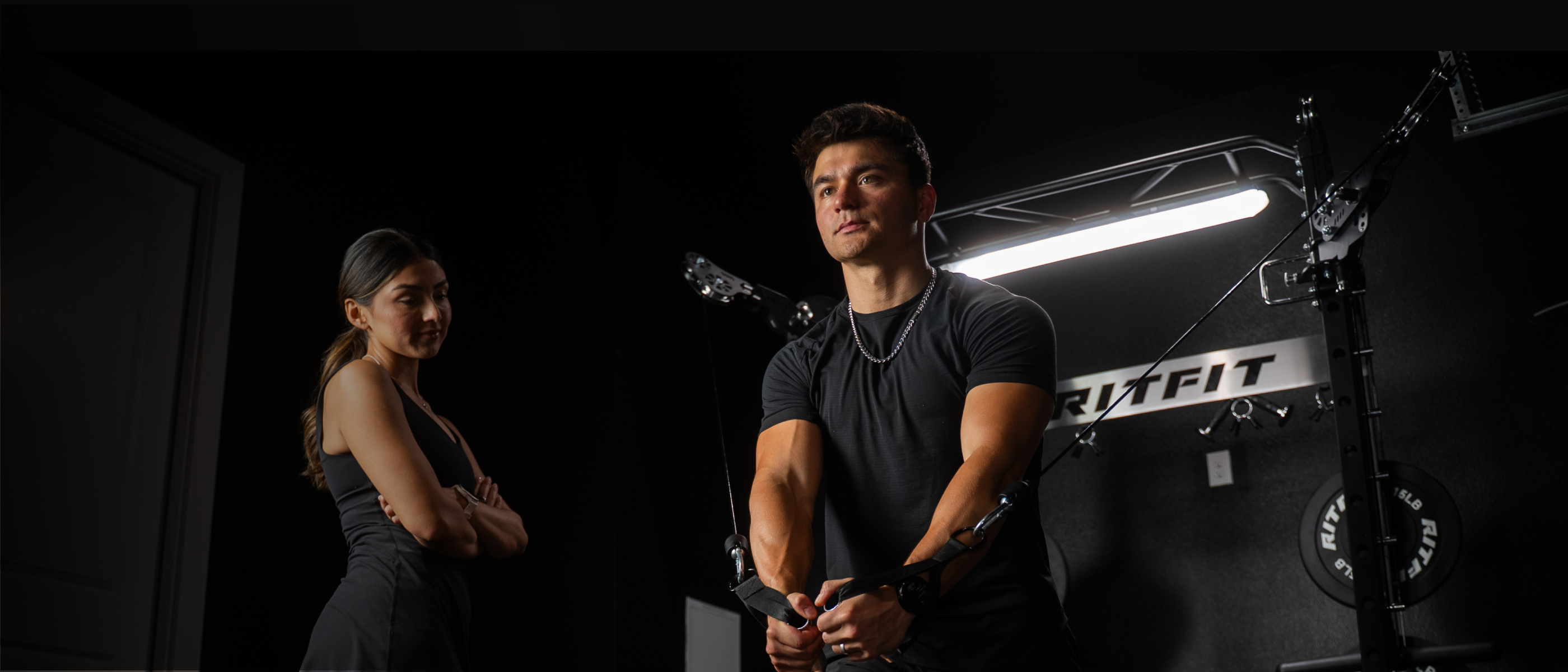 Man exercising on cable machine in dark gym with woman standing in background