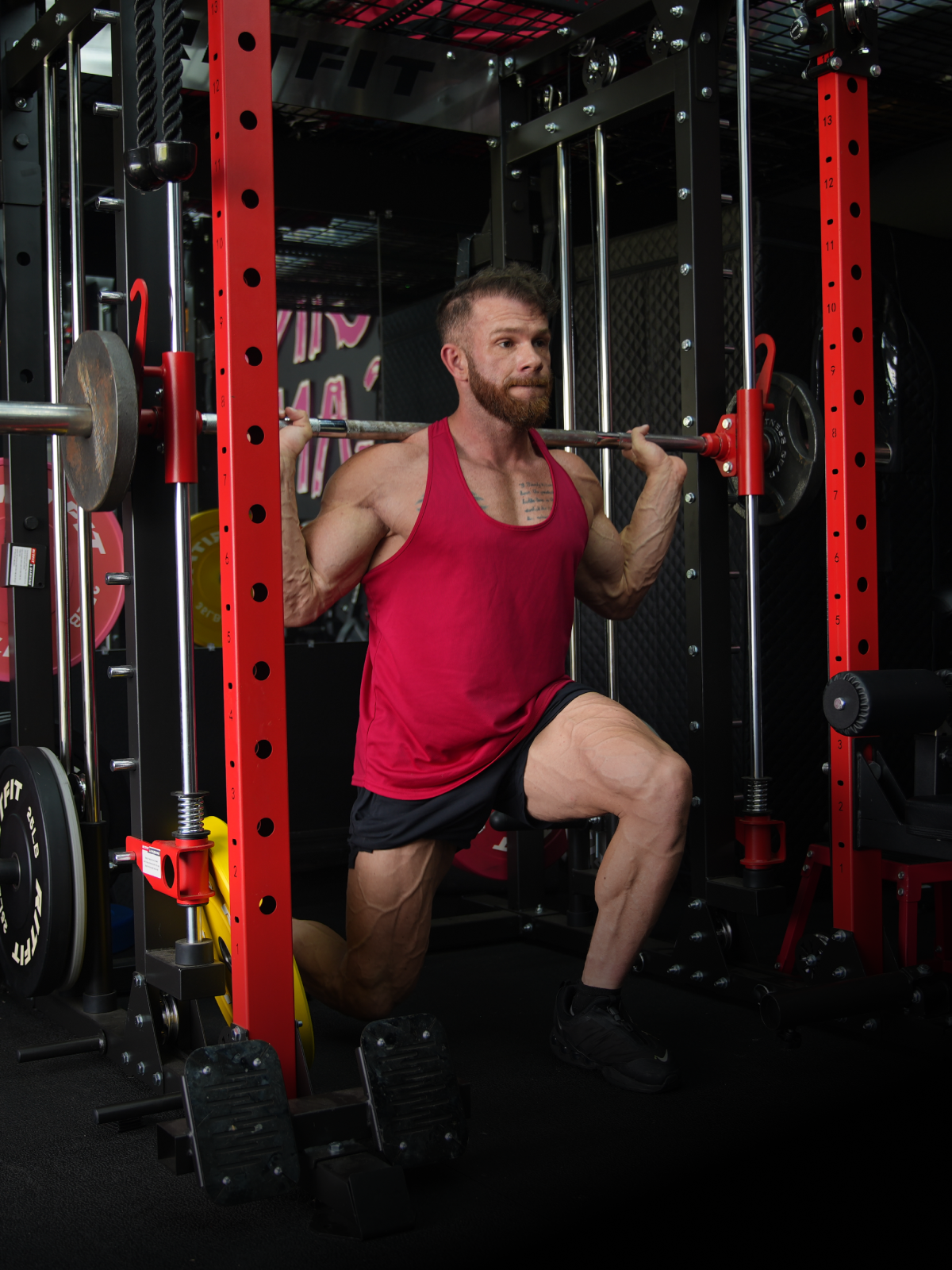 Man performing barbell lunge in red power rack wearing red tank top and black shorts