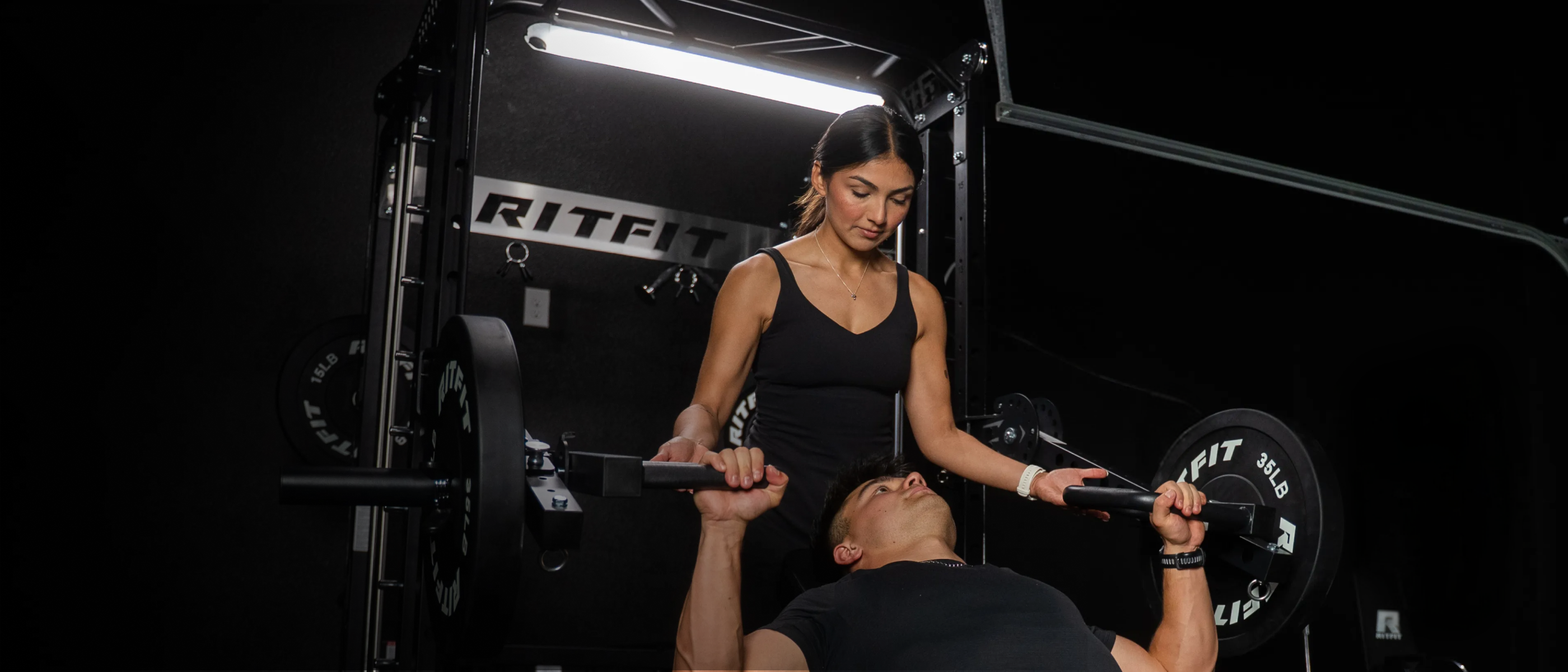 Woman spotting man bench pressing on black RitFit power rack